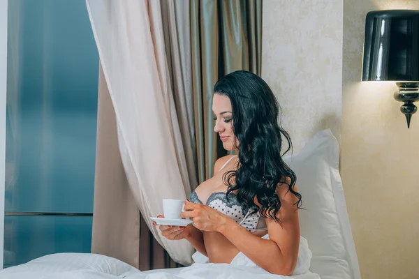 Porträt einer schönen jungen Frau in Nachtwäsche mit Kaffeetasse auf dem Bett. — Stockfoto