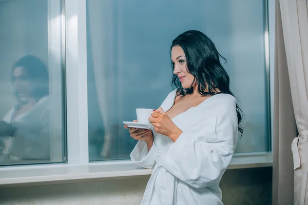 Caliente con café fresco. Hermosa joven en albornoz blanco bebiendo café y mirando a través de una ventana — Foto de Stock