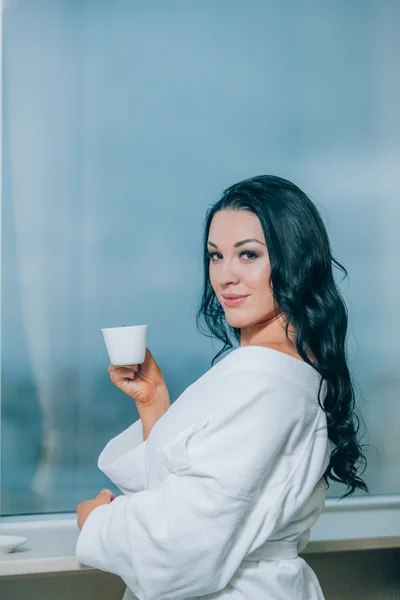 Getting warm with fresh coffee. Beautiful young woman in white bathrobe drinking coffee and looking through a window — Stock Photo, Image