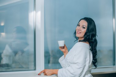 Taze kahve ile yaklaşıyor. Kahve içme ve bir pencereden bakarak beyaz bornoz güzel genç kadın