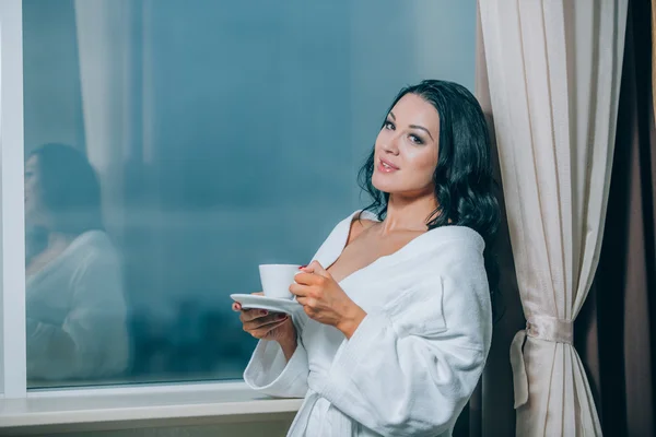 Getting warm with fresh coffee. Beautiful young woman in white bathrobe drinking coffee and looking through a window — Stock Photo, Image