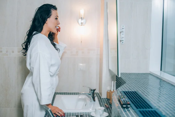 Portrait of attractive woman brushing teeth in bathroom  a white coat. healthy teeth. — Stock Photo, Image
