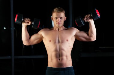 Handsome power athletic man with dumbbell confidently looking forward. Strong bodybuilder six pack, perfect abs, shoulders, biceps, triceps and chest