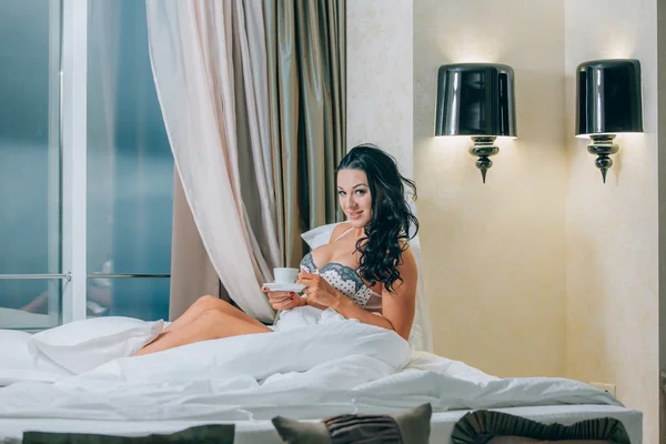 Portrait of beautiful young woman in nightwear holding coffee cup on bed. — Stock Photo, Image