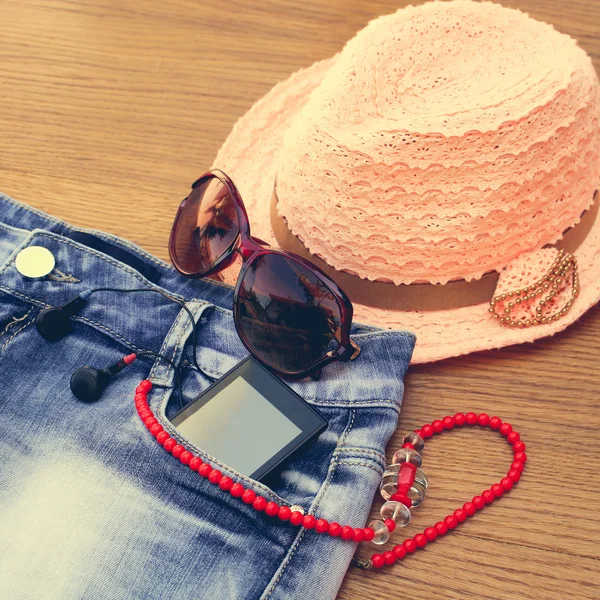 Summer women's accessories: red sunglasses, beads, denim shorts, mobile phone, headphones, a sun hat. Toned image