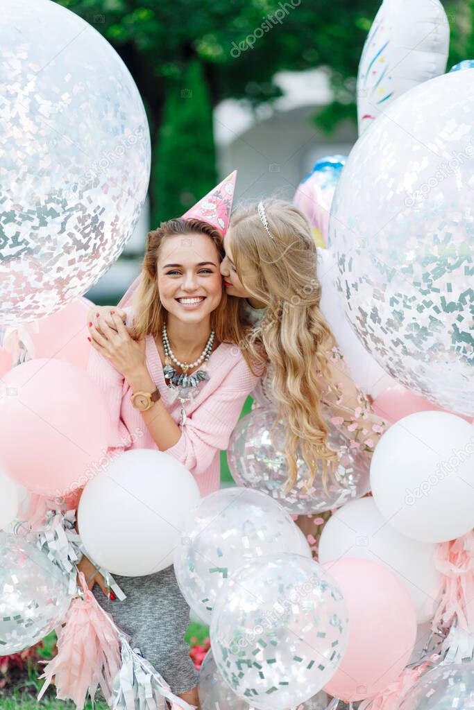 Two girls having fun in the park. They are in pink dresses and with a bunch of giant balloons. One kisses the other on the cheek