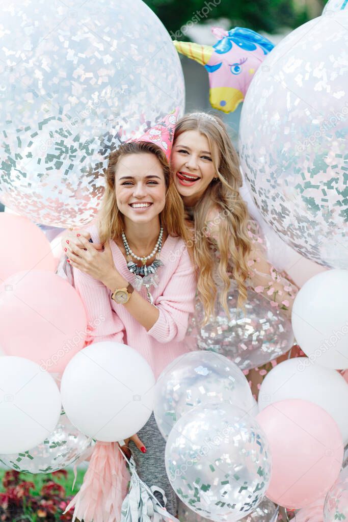 Two girls having fun in the park. They're wearing pink dresses and carrying a bunch of giant balloons.