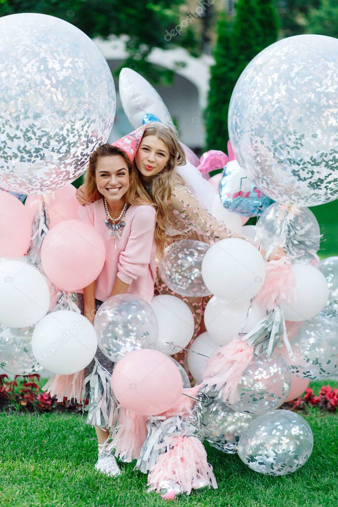 Two girls having fun in the park. They're wearing pink dresses and carrying a bunch of giant balloons.