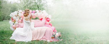 The bride is sitting on a bed in the spring in a park in a white boudoir dress. She stands by a tree with white flowers The bed is decorated with pink balloons