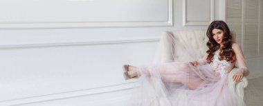 Beyaz iç çamaşırlı, güzel, yarı şeffaf elbiseli bir gelin. Kanepede oturuyor, çok güzel bir düğün saç stili ve makyajı var.