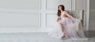 Beyaz iç çamaşırlı, güzel, yarı şeffaf elbiseli bir gelin. Kanepede oturuyor, çok güzel bir düğün saç stili ve makyajı var.