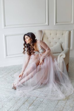 Beyaz iç çamaşırlı, güzel, yarı şeffaf elbiseli bir gelin. Kanepeye oturur ve ayakkabılarını giyer.