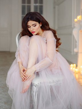 Beyaz iç çamaşırlı, güzel, yarı şeffaf elbiseli bir gelin. Sandalyeye oturuyor ve dizlerine sarılıyor. Arkasında şömine var.