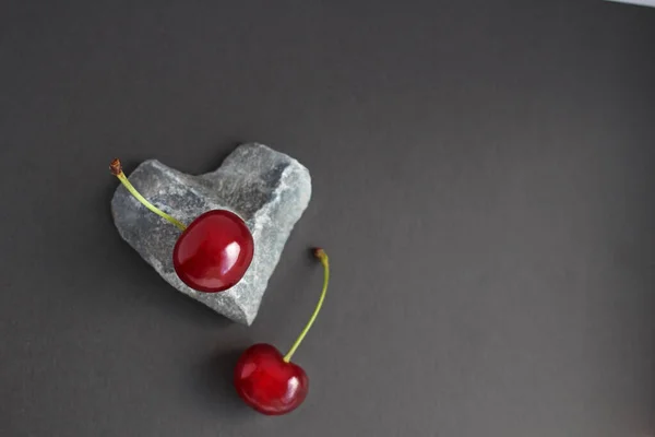 Cherry berries located on a stone in the form of a heart, and cherry leaves on a dark background — Stock Photo, Image
