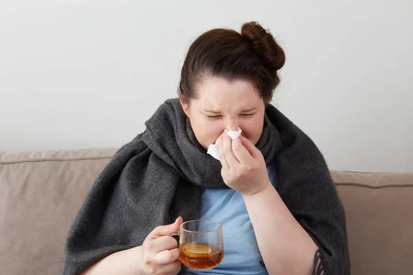 Una mujer joven está enferma con una taza de té caliente, una bufanda gris cálido alrededor de su cuello. — Foto de Stock