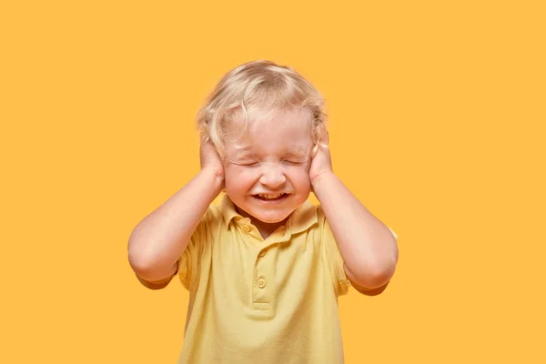 Ein fröhlicher netter blonder Junge lächelt und bedeckt seine Ohren mit seinen Händen. — Stockfoto