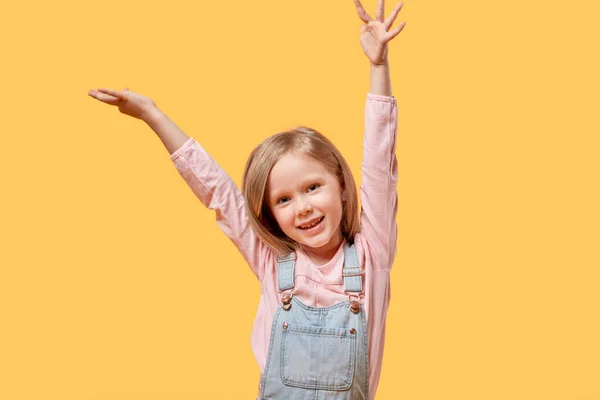 Das kleine blonde Mädchen hob glücklich die Hände in die Luft. Gelber Hintergrund. — Stockfoto