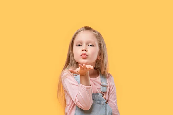 Ein blondes Mädchen blickt in die Kamera und gibt ihr einen Kuss. Gelber isolierter Hintergrund. — Stockfoto