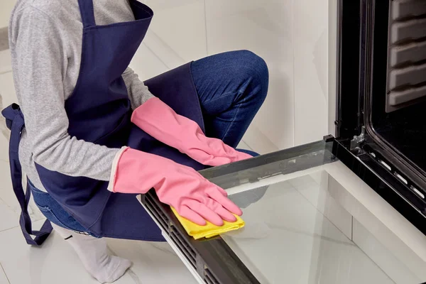 A woman washes the oven door with a microfiber cloth in rubber gloves.