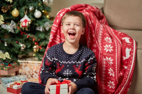 Um menino rindo bonito em uma camisola de rena segura um presente em sua mão. — Fotografia de Stock