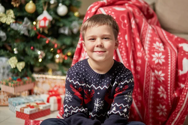 Rapaz bonito em uma camisola de rena olha para a câmera, ao lado da árvore de Natal. — Fotografia de Stock