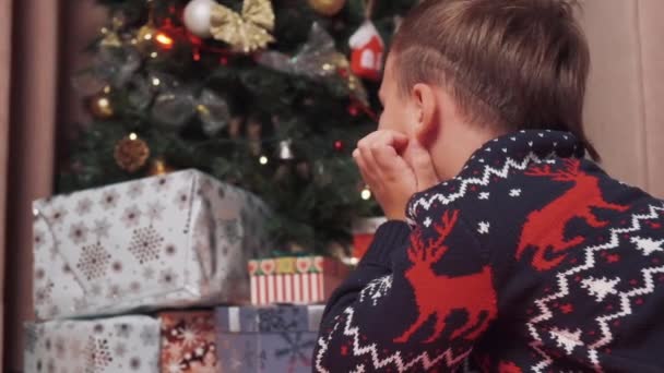 Мальчик дома лежит на полу и смотрит на елку и подарки. — стоковое видео