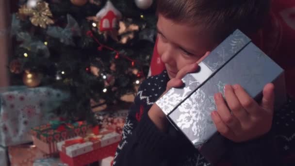 Un niño sorprendido sostiene y abre una caja de regalo, una luz azul brillante brilla dentro. — Vídeo de stock