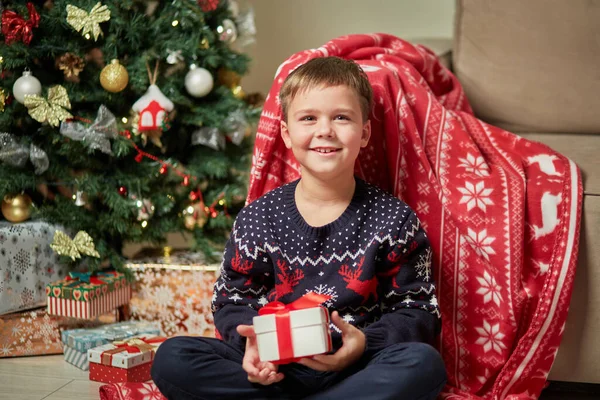 Rapaz bonito segurando um presente em sua mão, no fundo de uma árvore de Natal. — Fotografia de Stock