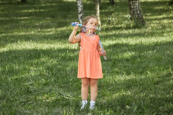 Uma menina em um vestido laranja brilhante sopra bolhas de sabão na natureza. — Fotografia de Stock