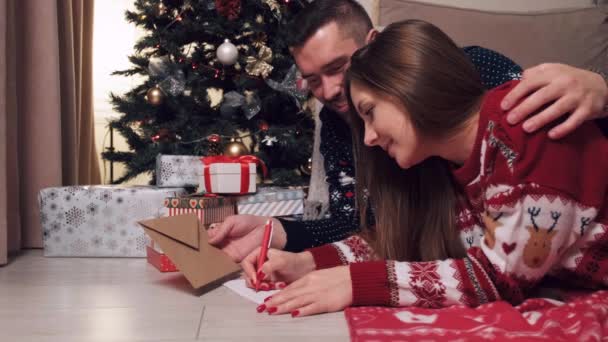 Un hombre y una mujer tumbados en el suelo escriben una carta a Santa Claus en papel. — Vídeo de stock