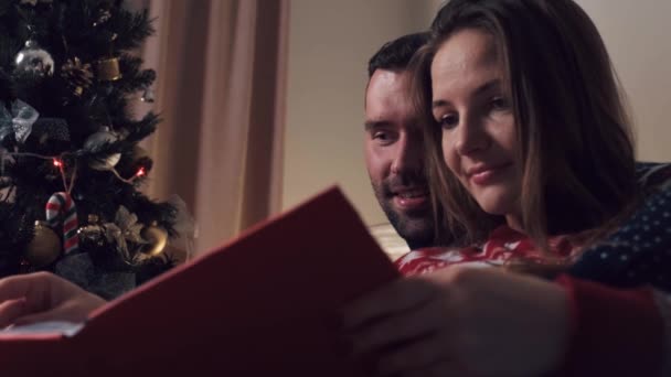 Hermosa pareja sentada en el sofá en Navidad mirando un álbum de fotos. — Vídeos de Stock