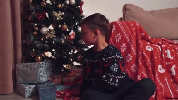 Το αγόρι κοιτάζει γύρω του και ανοίγει ένα χριστουγεννιάτικο δώρο, ένα φωτεινό μπλε λάμπει μέσα. — Αρχείο Βίντεο