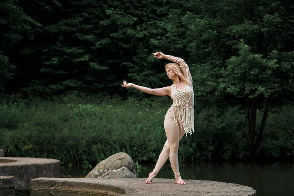 Une femme blonde danse pieds nus, avec un lac et un pont sinueux derrière elle. — Photo