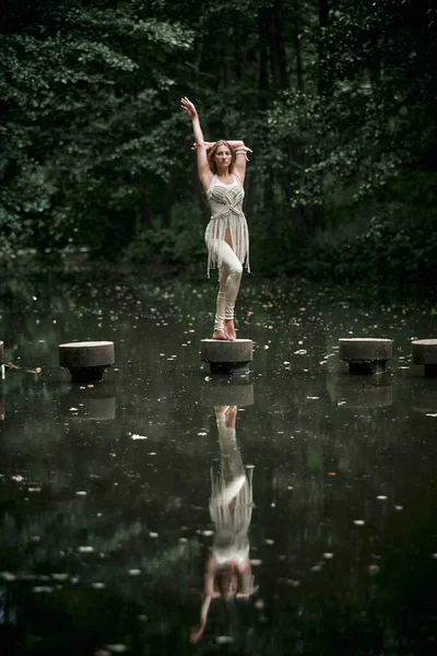 Yeşil bir ormanda, elbiseli genç bir kadın beton bir çemberin üzerinde duruyor..