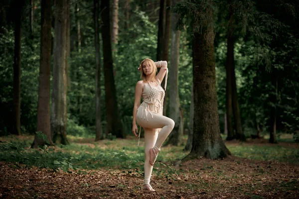 En ung blond kvinna står barfota på ett ben i en tät skog. — Stockfoto