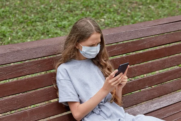 女の子は医療用のマスクと携帯電話を手にベンチに座っている. — ストック写真