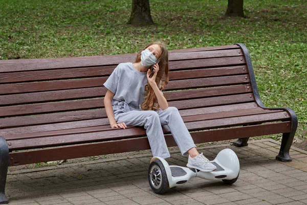 A menina está sentada em um banco com uma máscara médica e falando ao telefone. — Fotografia de Stock