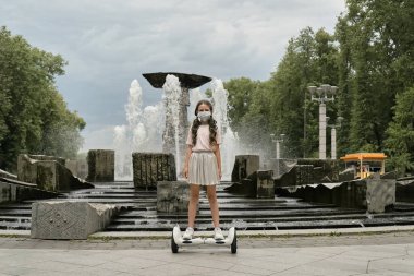 Etekli güzel bir kız, tıbbi maskeli bir parkta jiroskopun üzerinde duruyor..