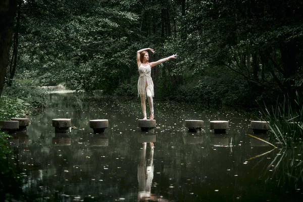 Vacker smal kvinna dansar på en betongbro nära en damm och skog. — Stockfoto