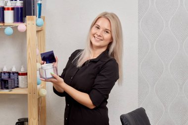Bir kuaför gülümser, elinde bir kavanoz krema tutar ve kameraya bakar..