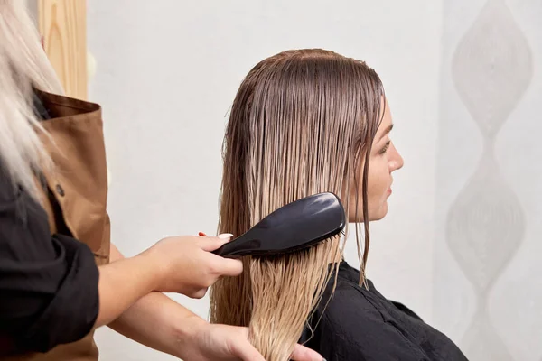 Um cabeleireiro feminino penteia os clientes cabelo loiro com um pente. — Fotografia de Stock