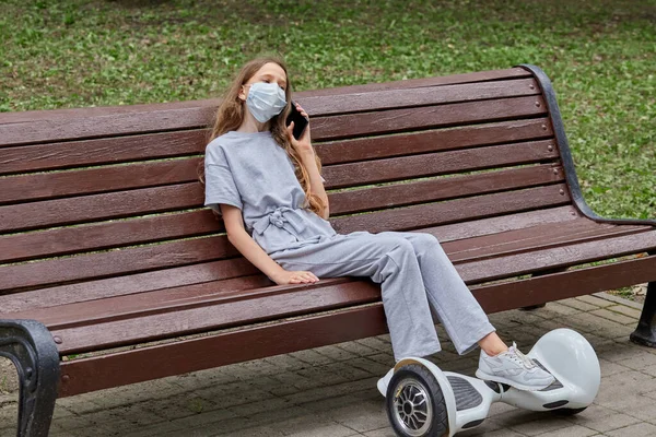 Uma menina com cabelos longos está sentada em um banco com uma máscara e falando ao telefone. — Fotografia de Stock