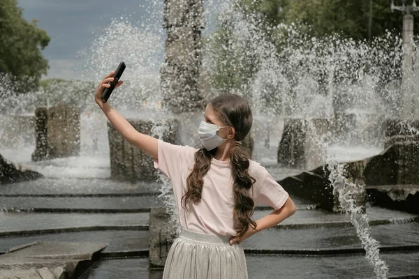 Uma adolescente com uma máscara médica em um parque tira uma selfie em seu smartphone. — Fotografia de Stock