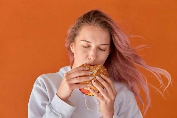 Mulher bonita bonito jovem com cabelo rosa e comer um hambúrguer. Fundo laranja — Fotografia de Stock