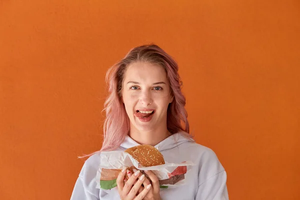 Mladá žena se podívá do kamery a olízne si rty. Držím velký hamburger.. — Stock fotografie