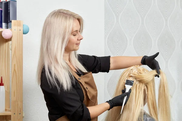 Een kapper in handschoenen schildert de haarwortels van een blonde klant met een borstel. — Stockfoto