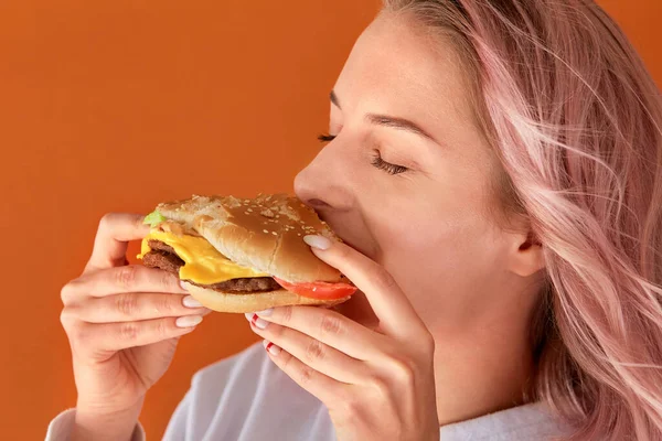 En ung kvinna med rosa hår äter snabbmat med njutning. Orange bakgrund. — Stockfoto
