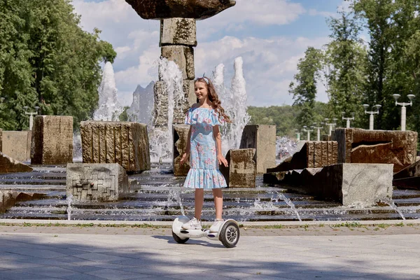Uma menina bonita em um vestido azul monta uma scooter giroscópio, prancha no verão. — Fotografia de Stock