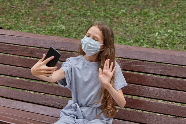 Una chica con una máscara médica se toma una selfie sentada en un banco en el parque. —  Fotos de Stock