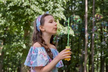 Omuzları açık bir elbise giyen bir kız parkta sabun köpüğü üflüyor..